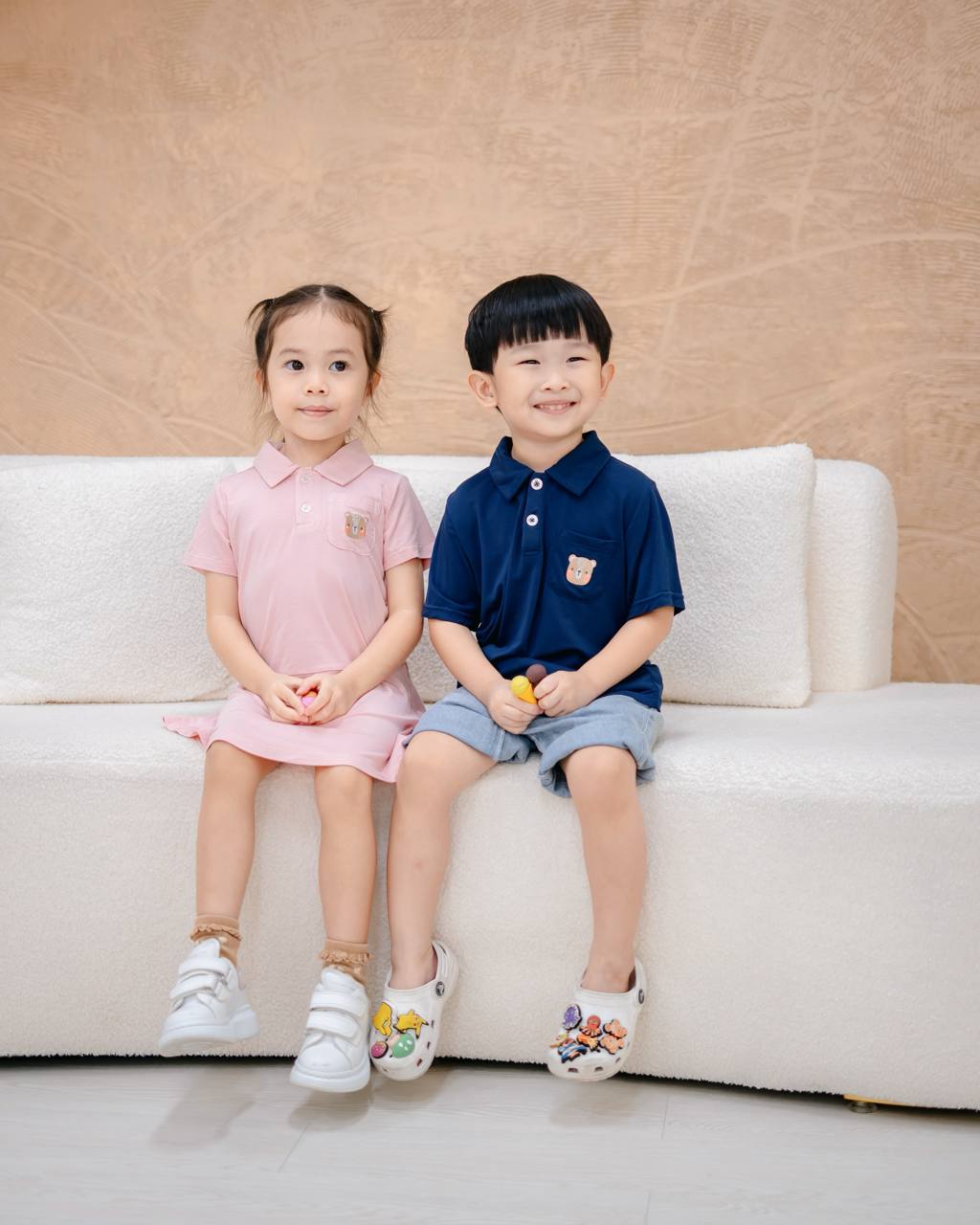 Two children sitting on a white couch against a beige wall.