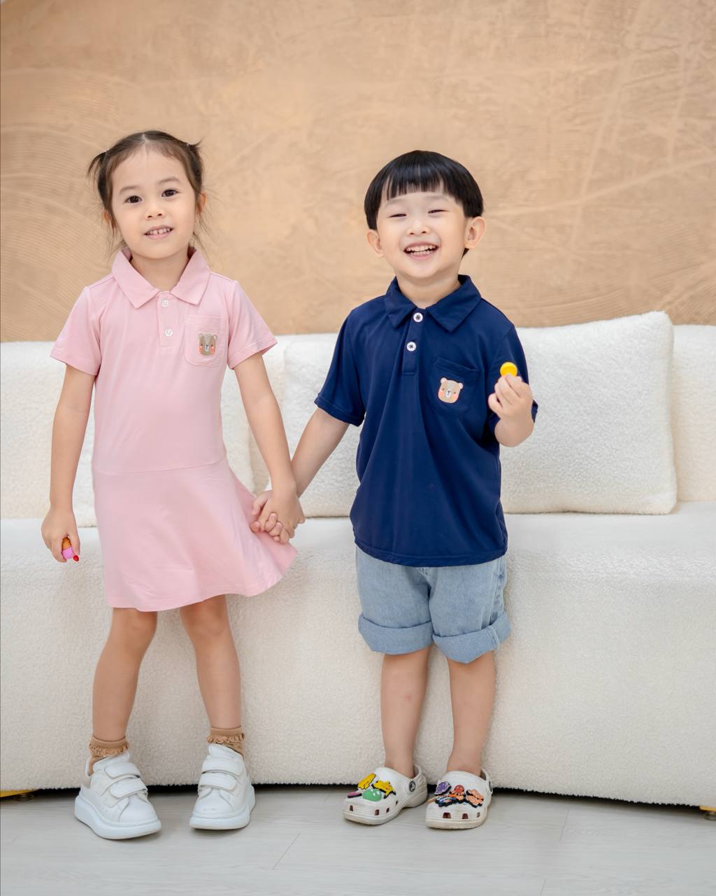 Two children holding hands, one in a pink dress and the other in a navy blue shirt with denim shorts, standing on a light-colored floor.