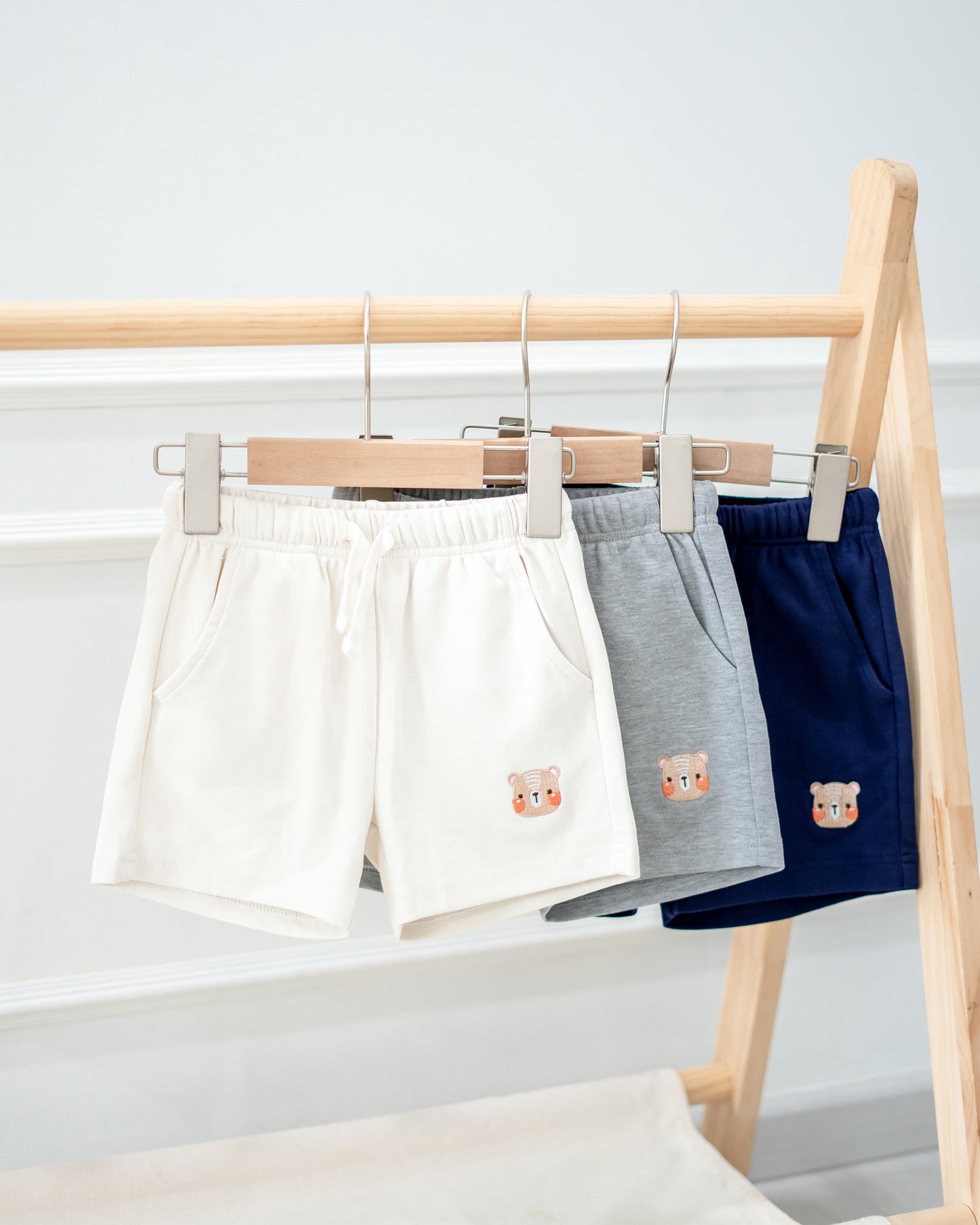LeBear Kids bear embroidered shorts in beige, grey, and navy displayed on wooden rack.