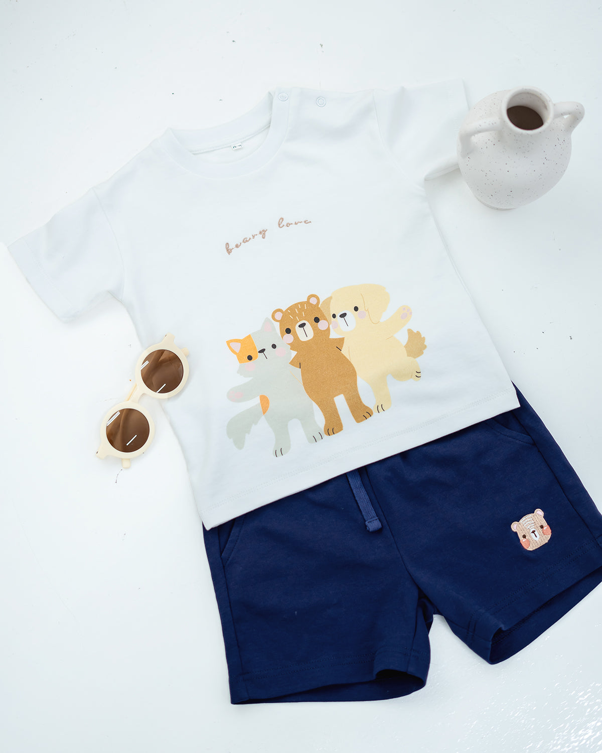 LeBear Kids Bear Friends Tee in white paired with navy bear embroidered shorts, flat lay with sunglasses and vase