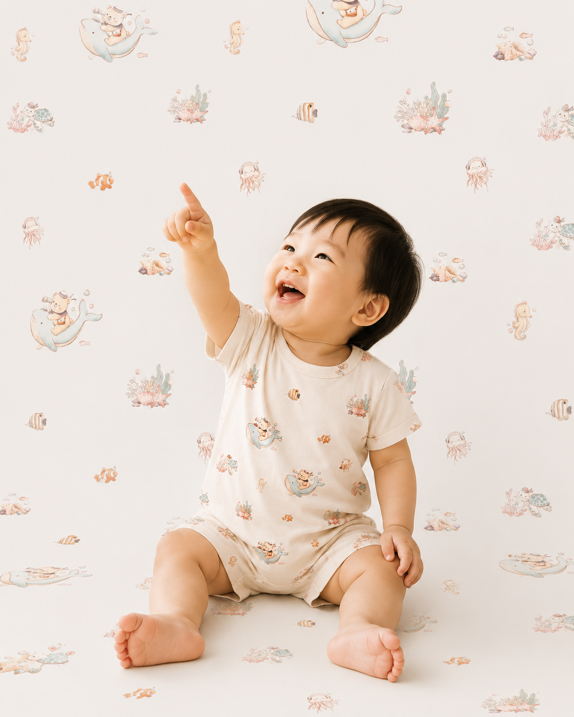 East Asian toddler wearing LeBear Ocean Dreams Tencel™ Onesie in Seashell Sand with ocean-themed print, sitting and pointing upwards against a clean studio background.