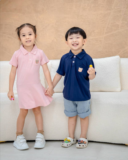 Two children holding hands, one in a pink dress and the other in a navy blue shirt with denim shorts, standing on a light-colored floor.