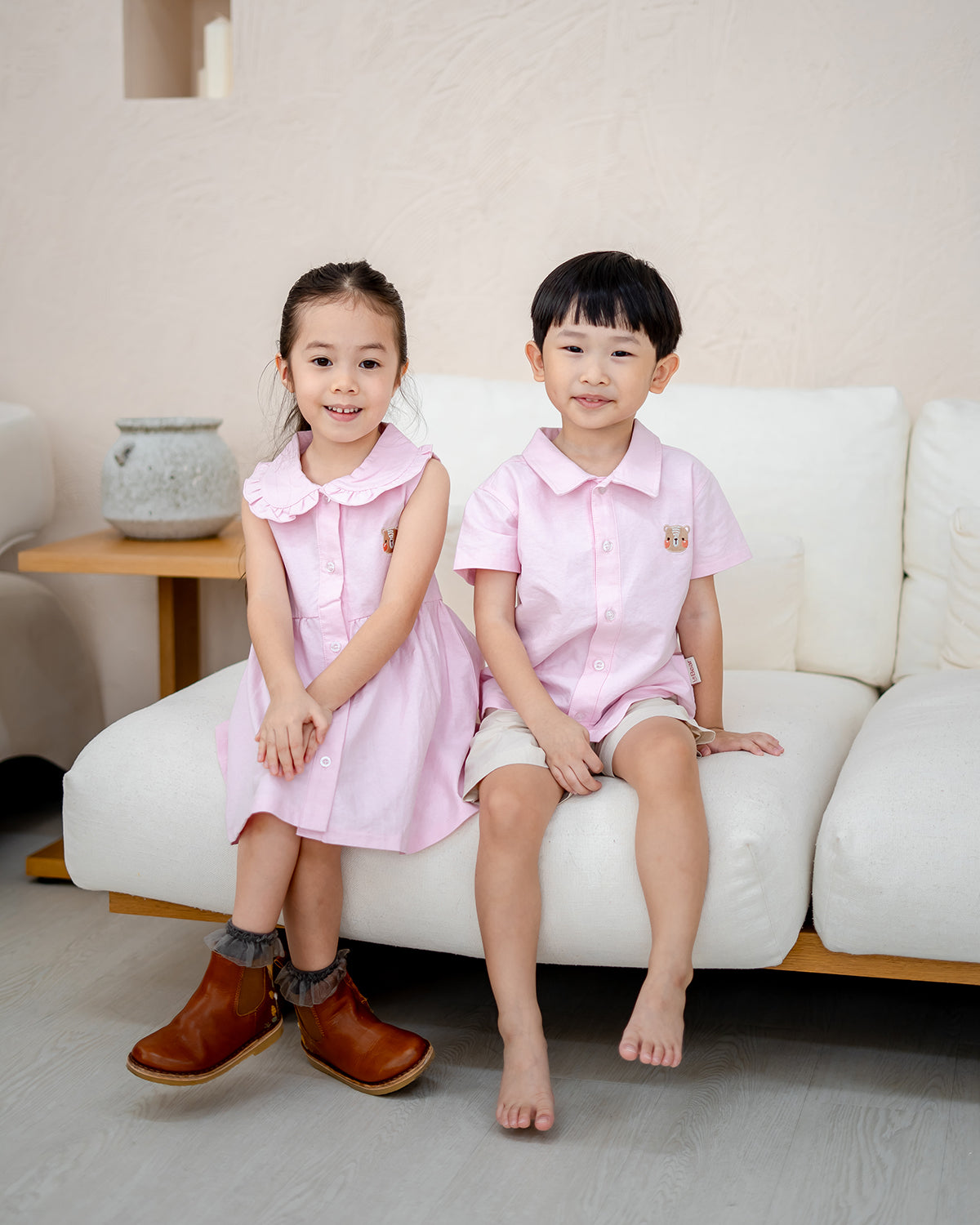 Girl in LeBear Atelier Dress and boy in matching Atelier Shirt in pink, sitting together on a sofa
