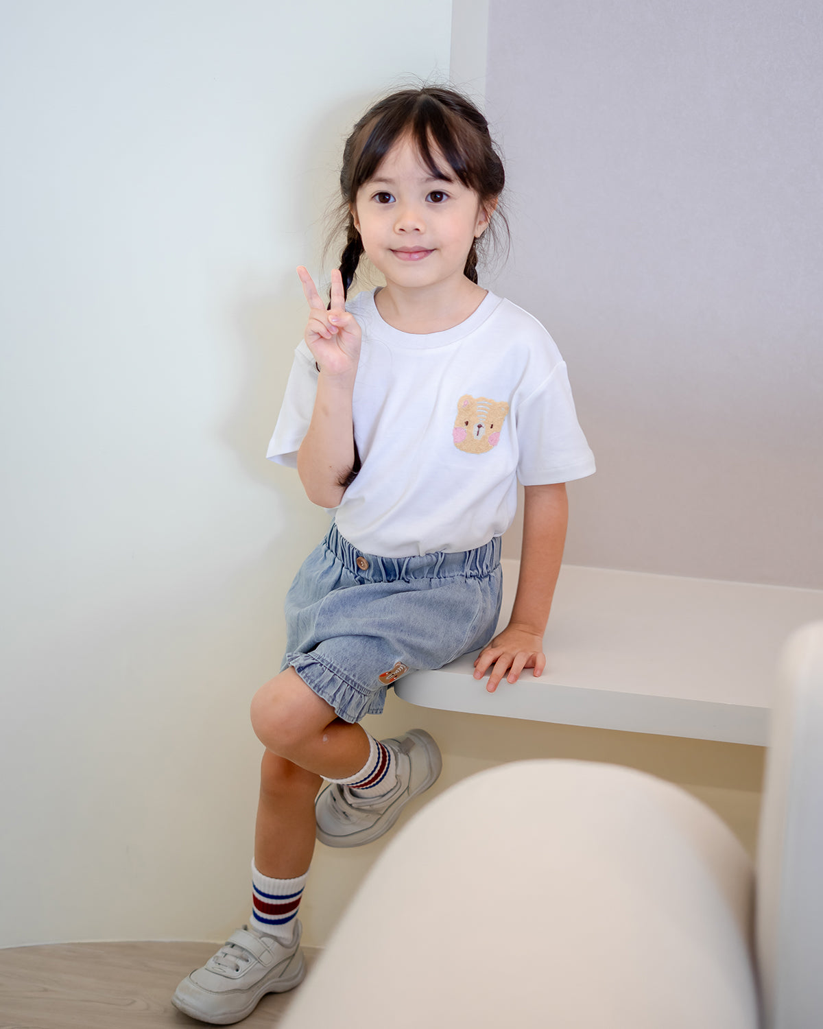 Little girl wearing LeBear Fluffy Embroidery Kids Tee in white premium cotton with denim ruffle shorts, posing with a peace sign – cute kids outfit by LeBear Singapore.