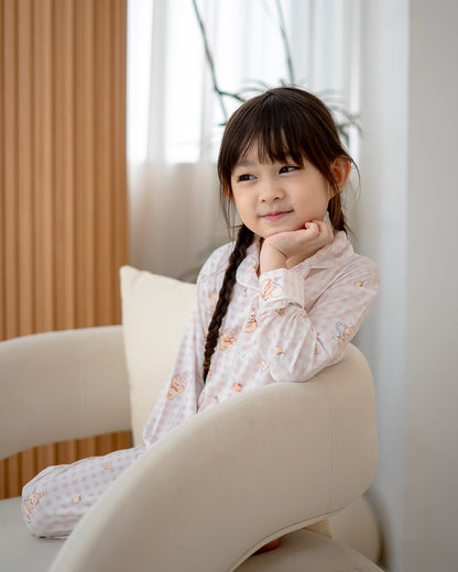 Girl wearing LeBear Ice Cream Bear pink gingham bamboo kids pyjamas sitting on armchair in bright home studio