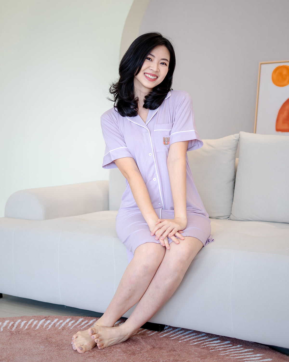 Woman in lilac LeBear bamboo pyjama dress with white piping and fluffy bear embroidery, seated comfortably on sofa – premium women’s loungewear by LeBear Singapore.