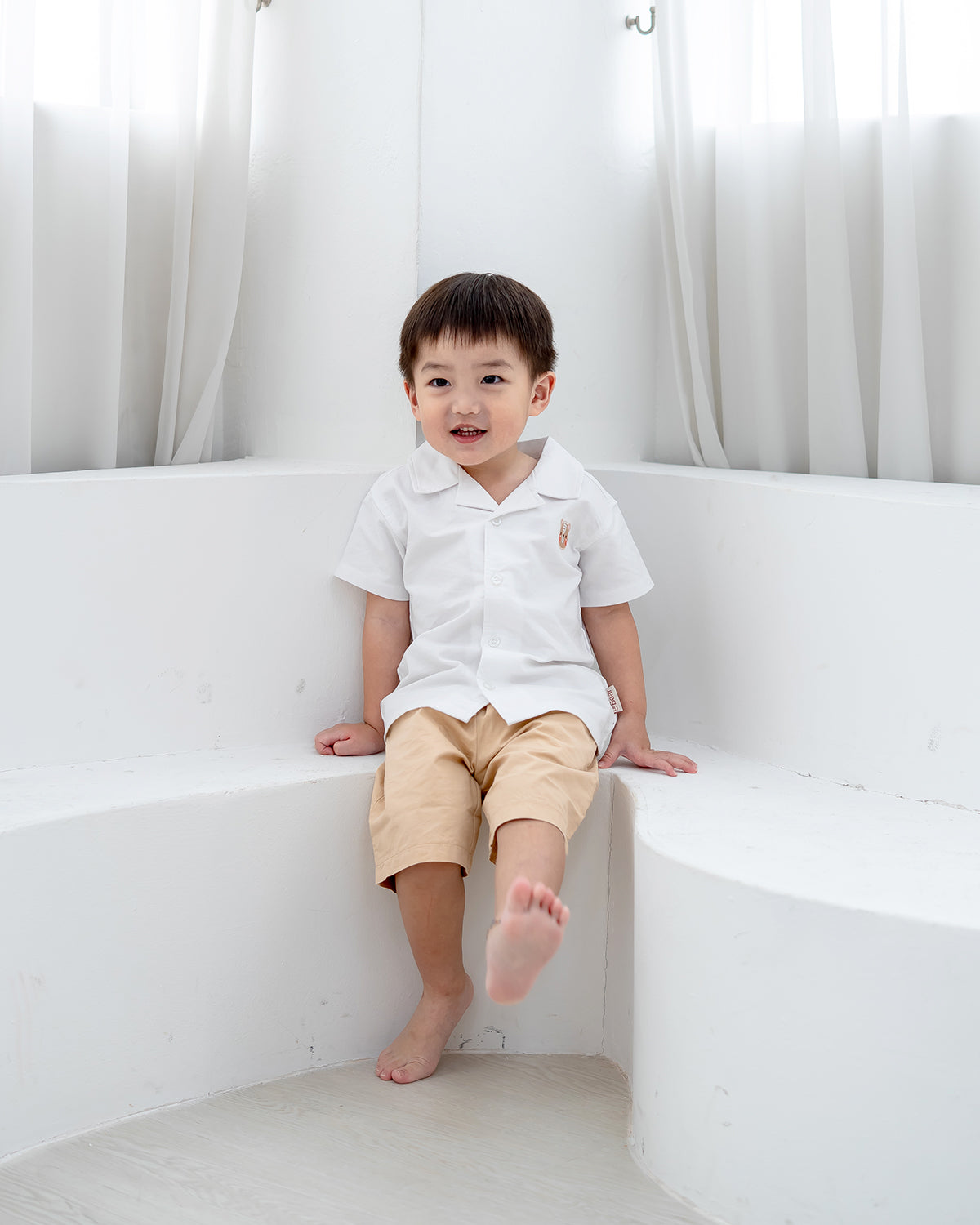 Boy in white camp collar shirt from LeBear sitting against white backdrop