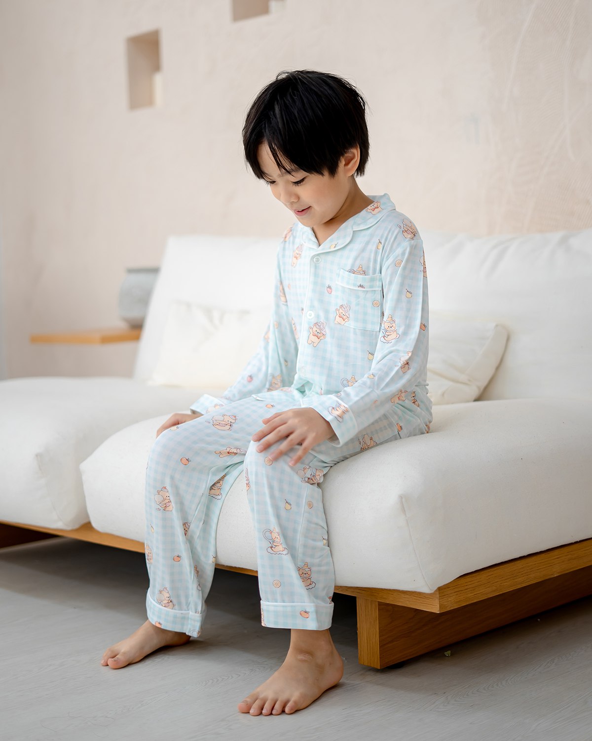 Boy wearing LeBear Sweet Scoops Ice Cream Bear blue gingham bamboo pyjamas sitting on bed in bright bedroom