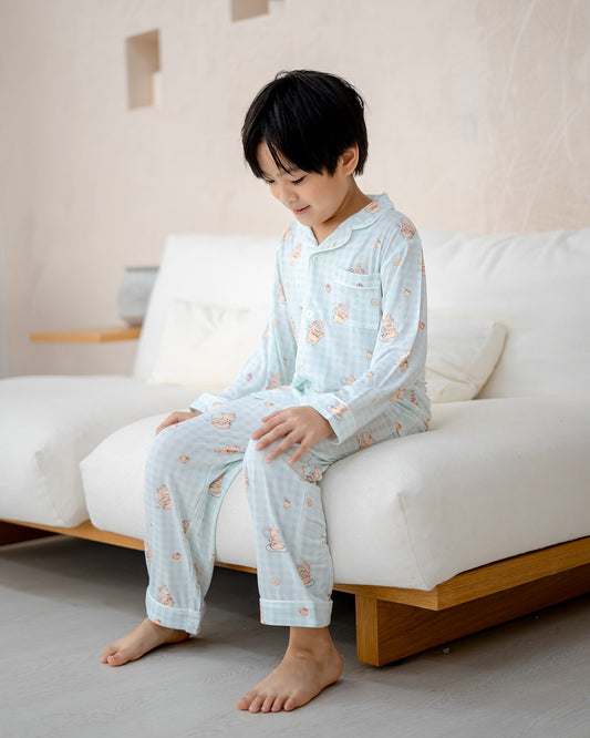Boy wearing LeBear Sweet Scoops Ice Cream Bear blue gingham bamboo pyjamas sitting on bed in bright bedroom